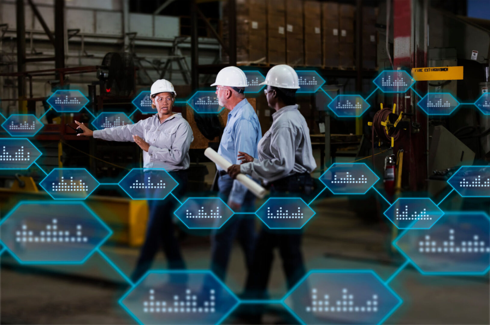 A multiracial group of three workers wearing hardhats and safety glasses, two mature women and a senior man, conversing in warehouse of a metal fabrication plant. The woman in charge, holding a walkie-talkie, is pointing at something, telling them what to do.