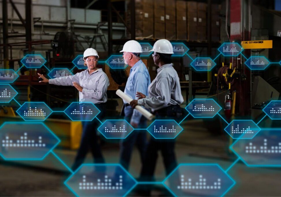A multiracial group of three workers wearing hardhats and safety glasses, two mature women and a senior man, conversing in warehouse of a metal fabrication plant. The woman in charge, holding a walkie-talkie, is pointing at something, telling them what to do.